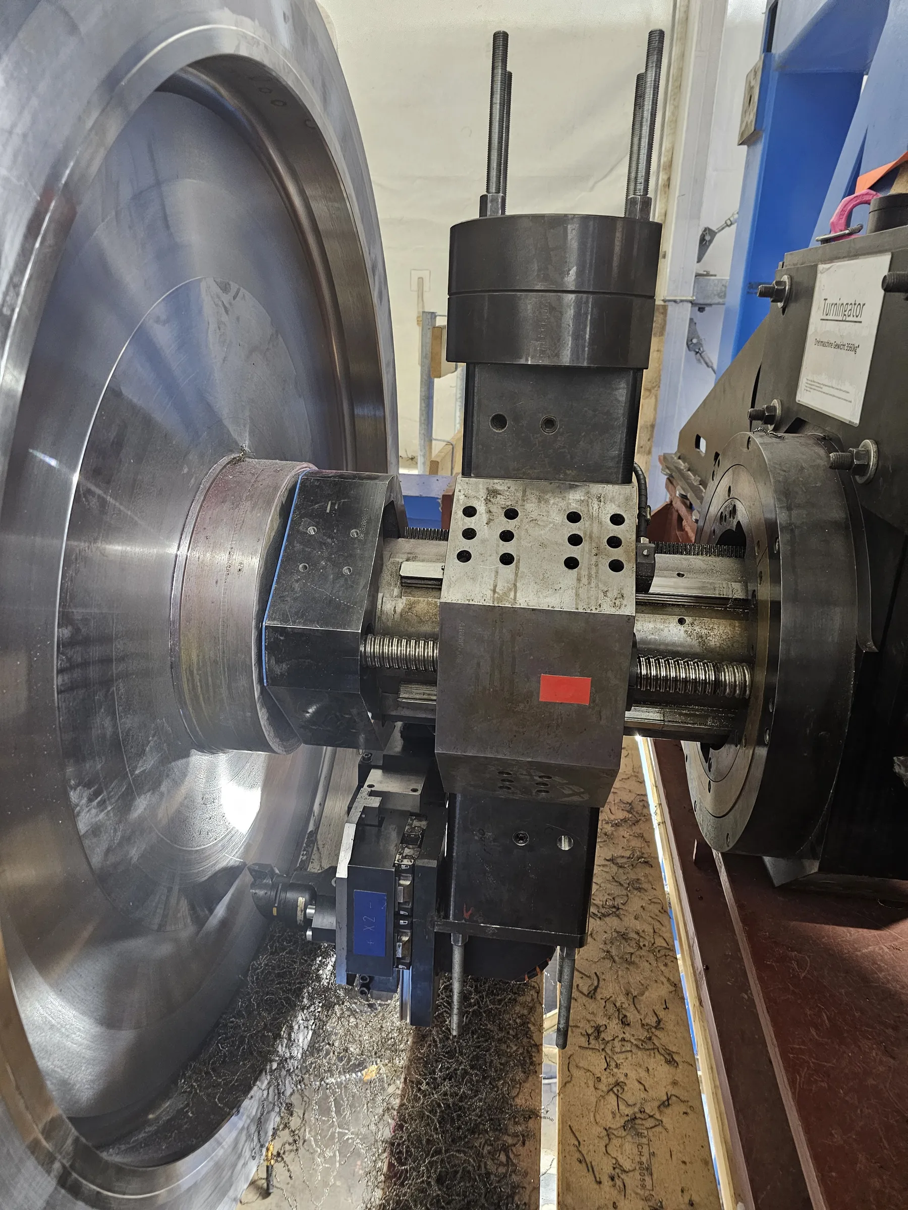 A large, circular, silver-colored metal workpiece is being machined by an industrial turning machine, generating numerous dark, curly metal shavings. The central machining mechanism consists of dark grey and black metal components, including a visible threaded rod. A cutting tool is actively engaged with the workpiece. On the right, a robust blue metal frame supports the machine, featuring a white sign that reads "Turningator" and "Drehmaschine Gewicht 3560kg". The floor below is covered with metal shavings and wooden planks. Bright overhead lighting illuminates the scene, creating reflections on the shiny metal surfaces.