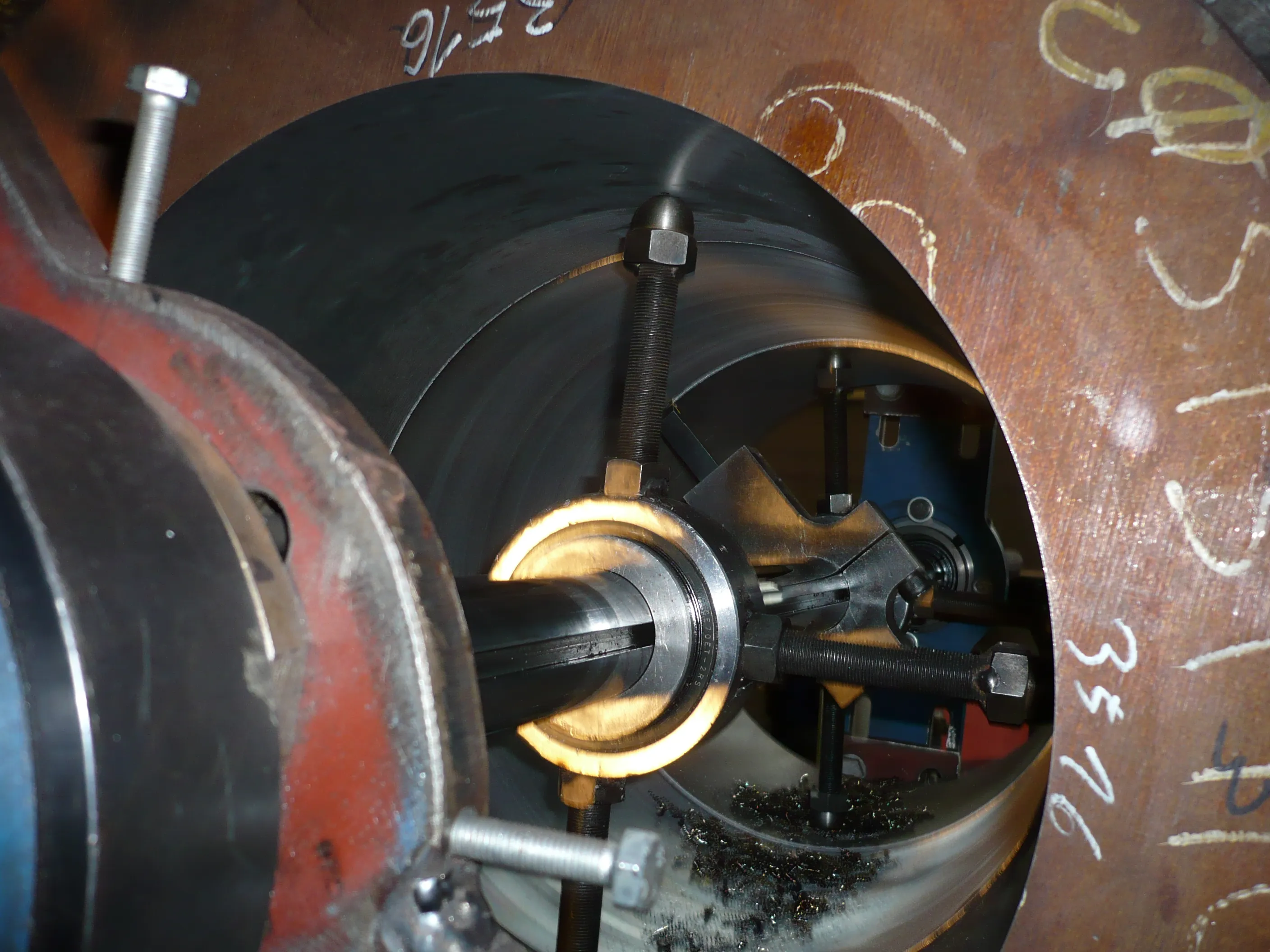 A close-up, well-lit industrial image captures a machining tool operating inside a large metal pipe. In the foreground, a red-painted machine part with a silver bolt is visible. The central focus is a metal shaft extending into a cutting tool assembly, actively producing dark metal shavings on the pipe's inner, dark grey surface. The pipe's outer, rusty brown surface displays white chalk markings, including "3516". Bright light illuminates the metallic components and the cutting area.