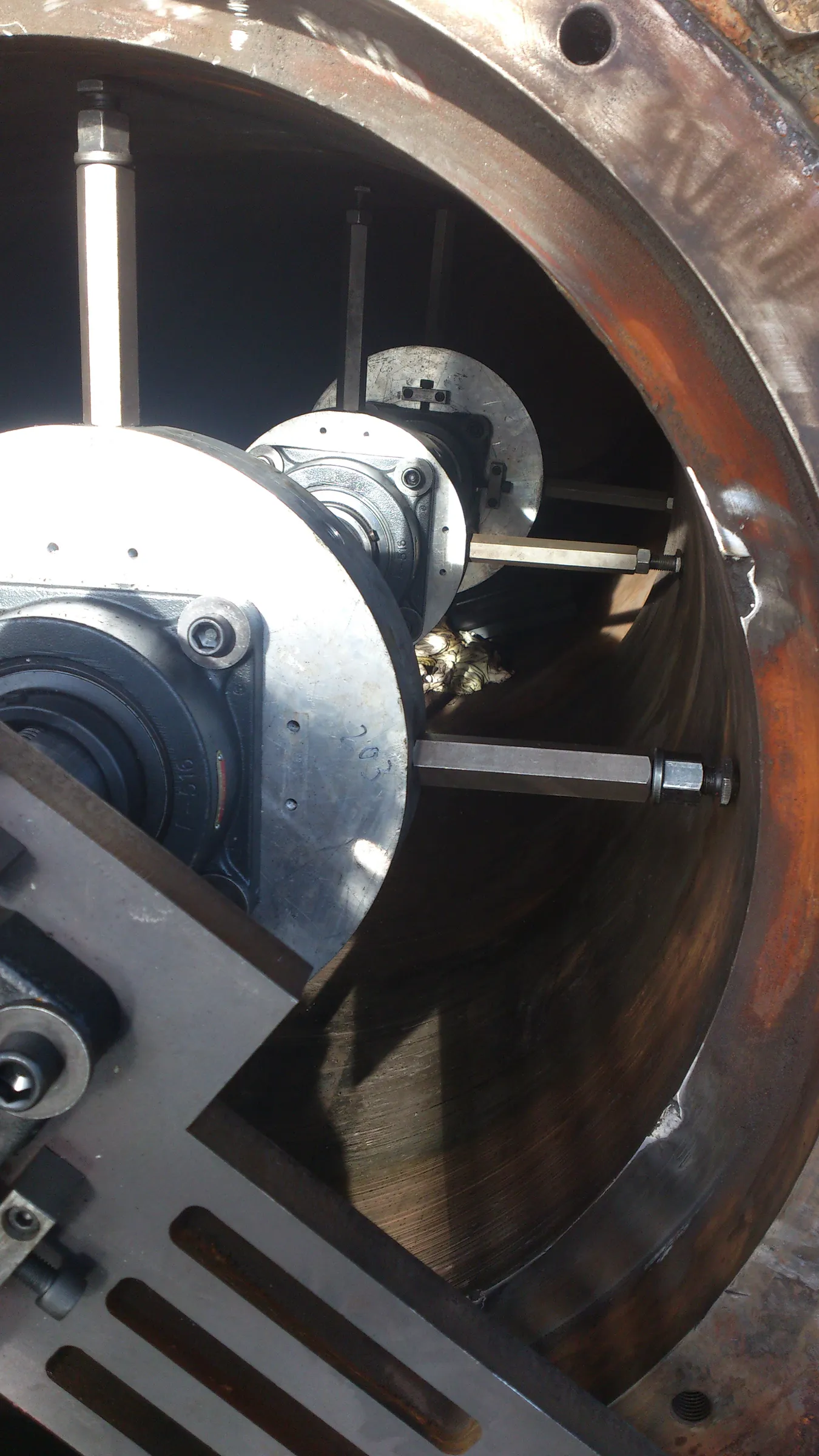 An angled view inside a large, rusty metal pipe reveals industrial machinery. Two circular metal components with black bearings are prominent, secured by hexagonal rods braced against the pipe's dark, oxidized interior. Bright light illuminates the machinery from the upper left, contrasting with the pipe's shadowed depths.