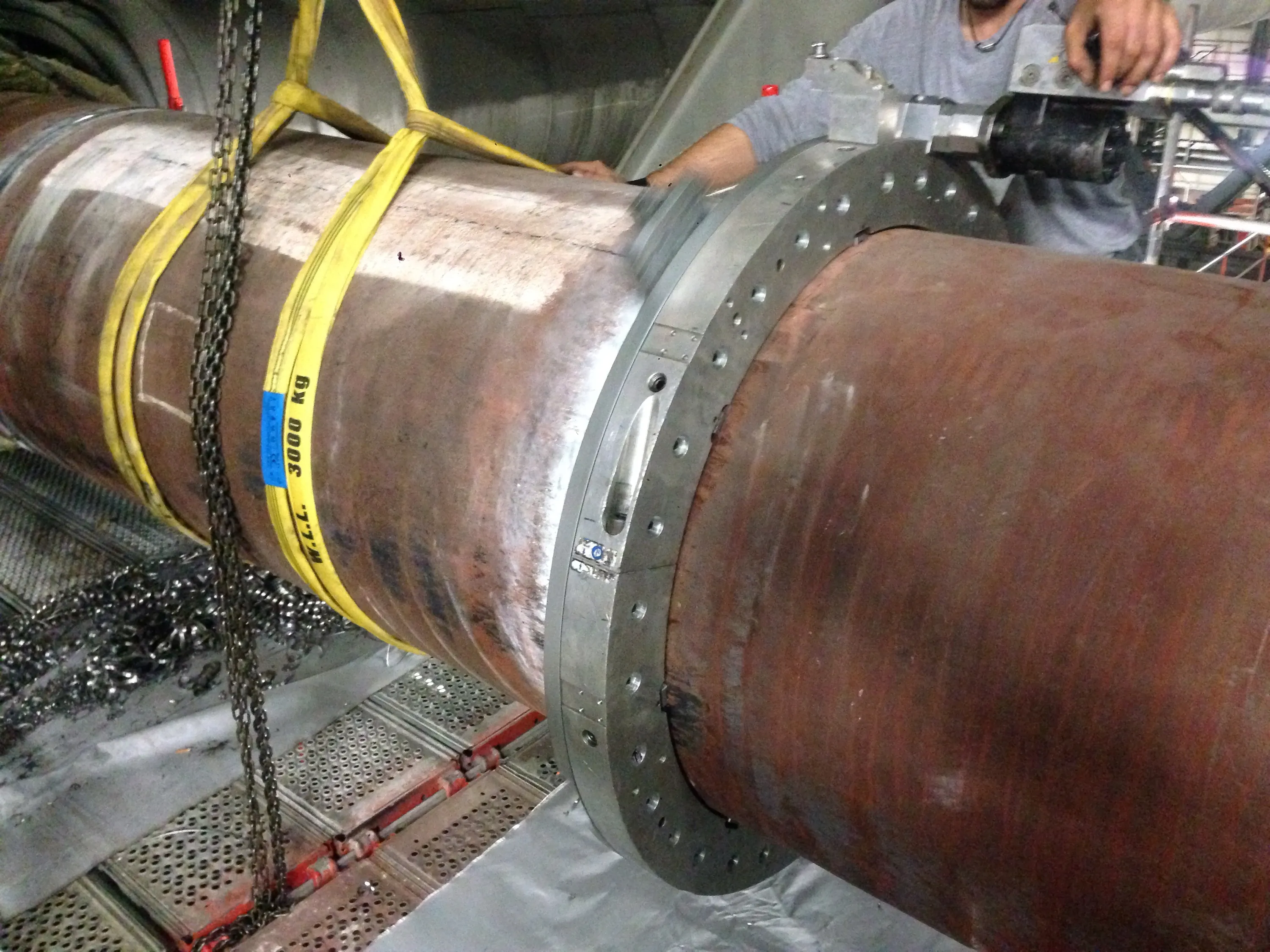 A large, rusty industrial pipe is suspended by bright yellow lifting slings and dark metal chains in an industrial setting. A worker in a grey shirt operates a machining tool attached to a silver flange on the pipe. Metal shavings are scattered on the perforated metal grating below, and the pipe shows areas of cleaned metal alongside its reddish-brown, oxidized surface.