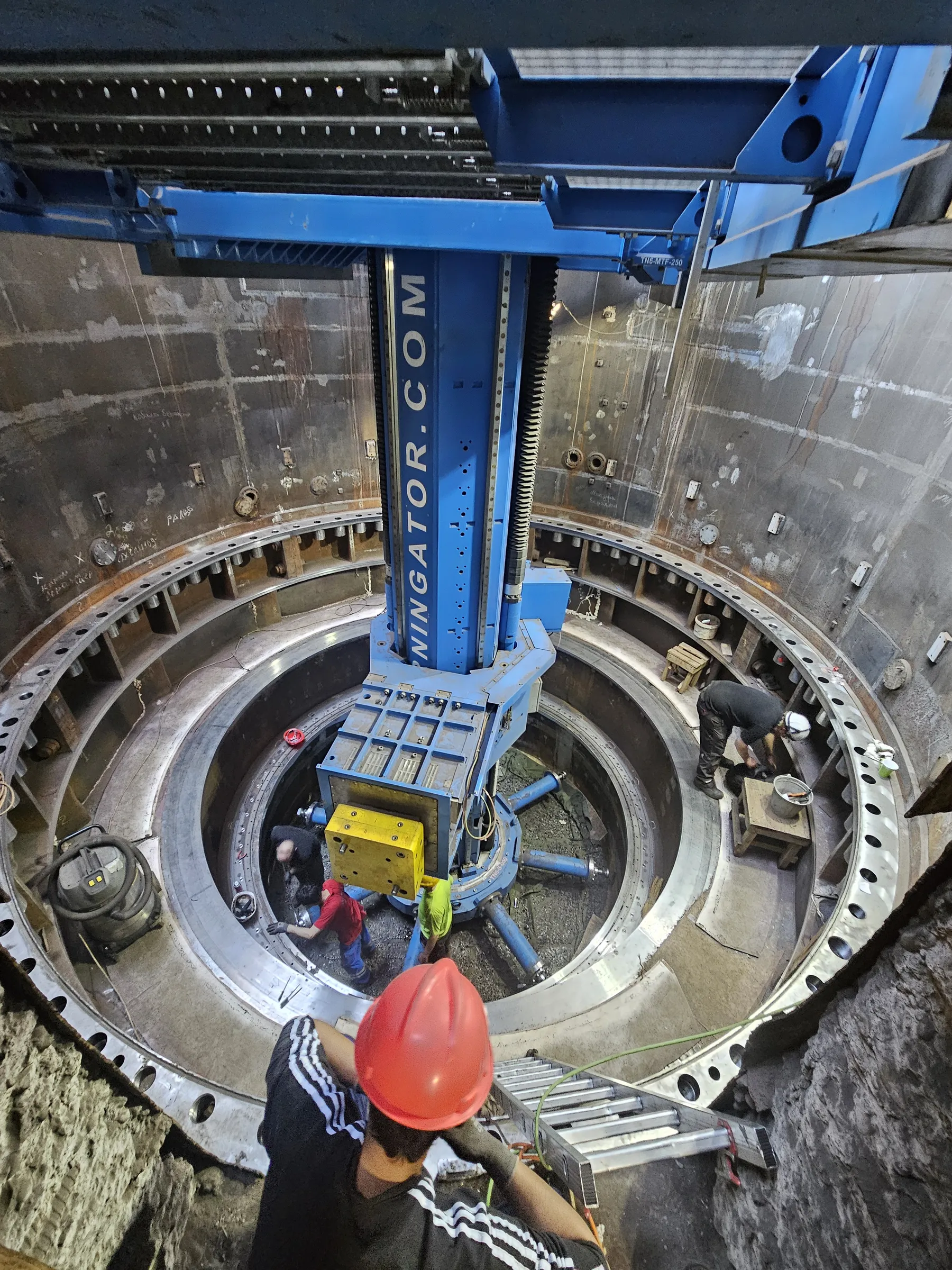 A deep, circular industrial pit features a massive blue boring machine, labeled 'NINGATOR.COM', positioned centrally. Multiple concentric, heavy metal rings with bolt holes line the pit's lower levels. Several workers, including one in a red hard hat in the foreground looking down from the pit's edge, are actively working around the machine. A silver ladder leans against the rough concrete wall in the foreground. The entire scene is brightly illuminated by artificial light.