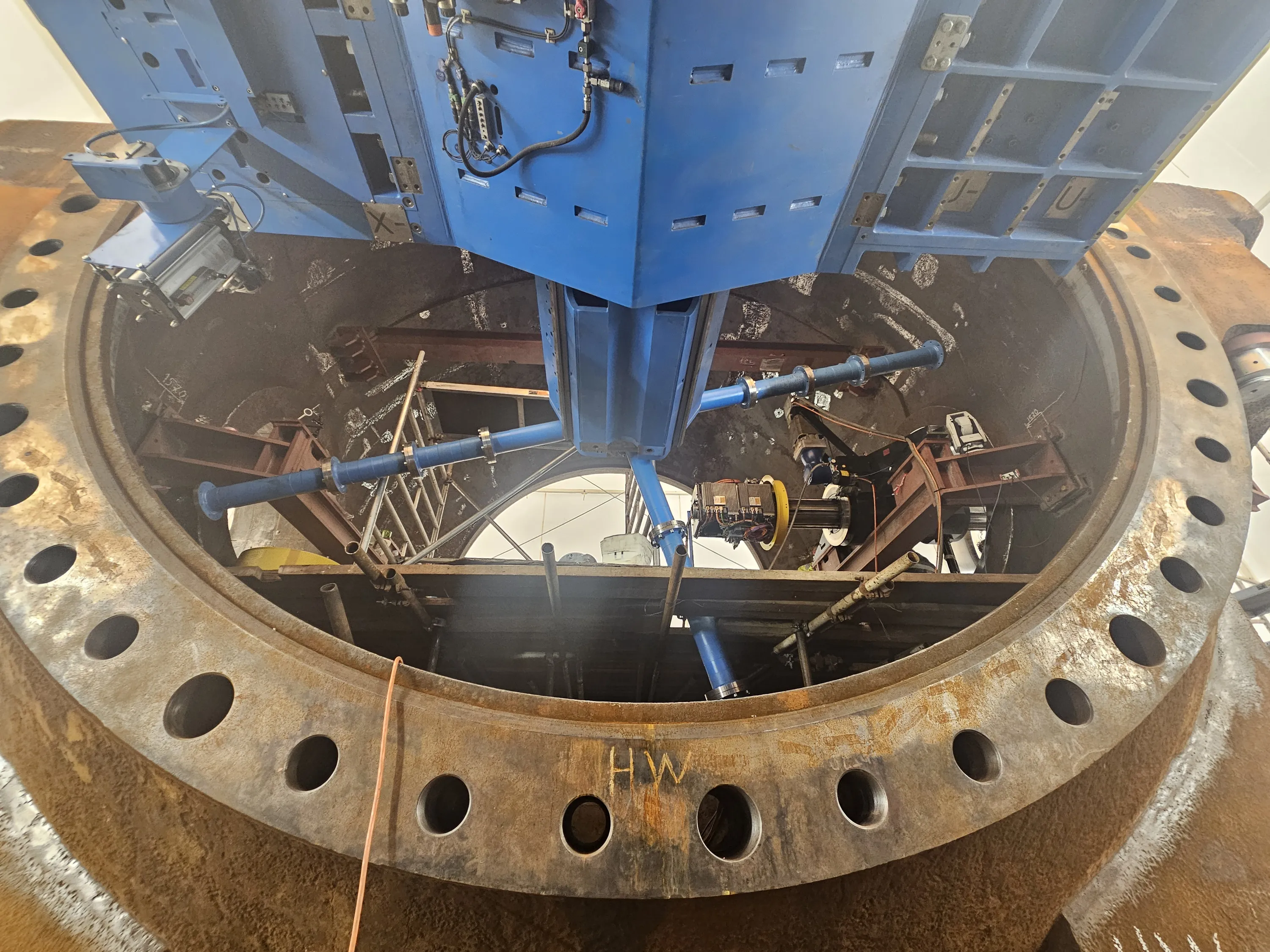 A top-down view shows a large, rusty metal flange with multiple bolt holes, marked "HW", surrounding an industrial chamber. A blue machine extends from above into the opening, equipped with cables, sensors, and a motor. Inside the chamber, blue pipes, metal support structures, and additional machinery, including a yellow-banded device and surveying equipment, are visible under industrial lighting, with a bright opening in the background.