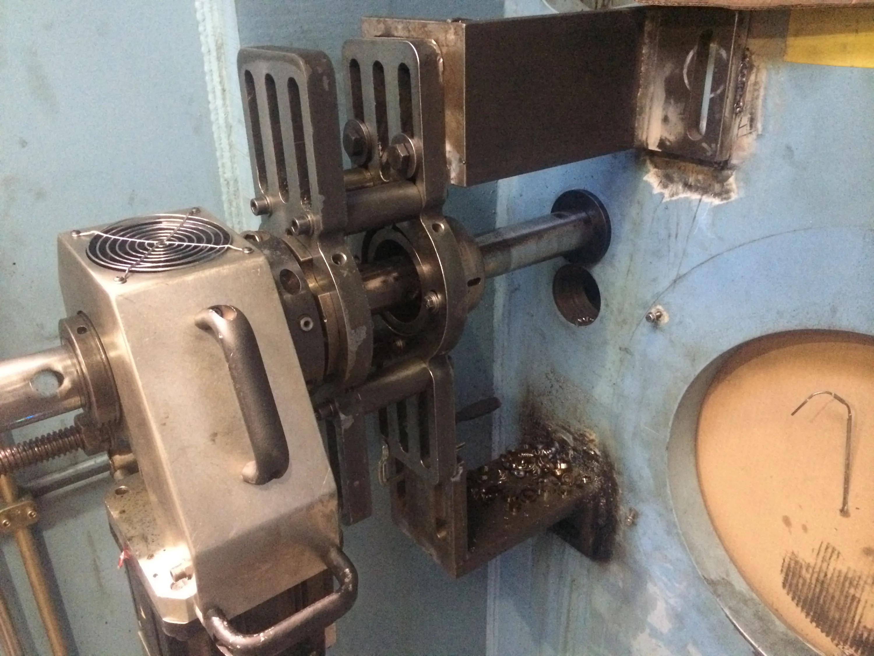 A detailed view of industrial machinery shows a central metal shaft passing through multiple grey metallic components, including a housing with a top-mounted cooling fan and handles. The shaft extends into a large light blue-grey machine panel, which features a circular opening revealing a tan interior. Accumulations of dark metal shavings are visible on a lower support bracket. The scene is brightly lit, emphasizing the functional metal textures.