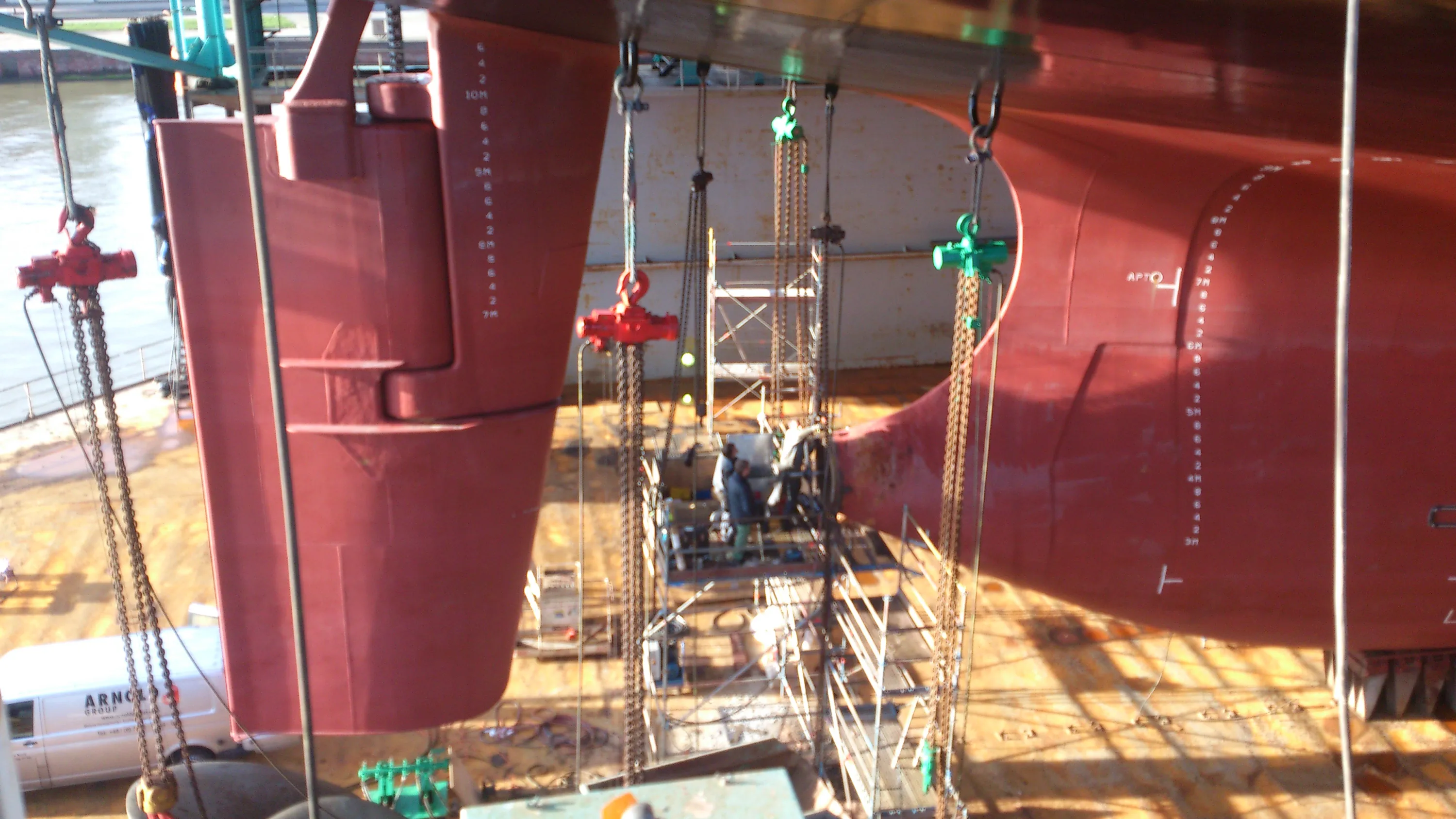 A large red ship hull, marked with white draft numbers, is seen in a dry dock. Red and green chain hoists hang from overhead structures, with workers on scaffolding near the hull. Bright sunlight illuminates the scene, casting sharp shadows on the brown ground where a white van is visible.