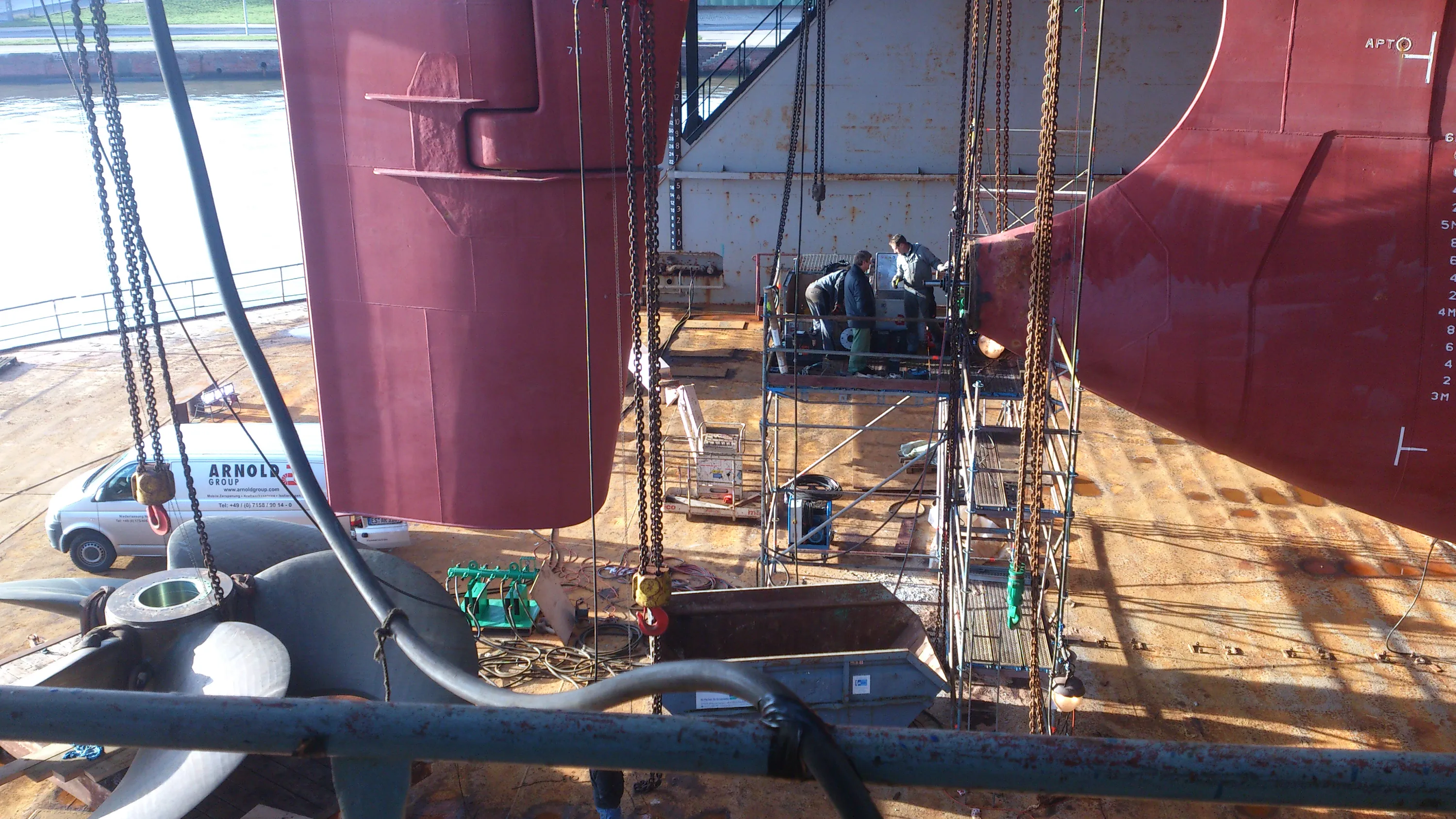 A large reddish-brown ship's stern and propeller are seen in a dry dock, with three workers on scaffolding near the hull. Heavy chains hang around the work area, and a white service van is parked on the rusty dry dock floor. Bright sunlight casts strong shadows, illuminating the industrial setting and background water.