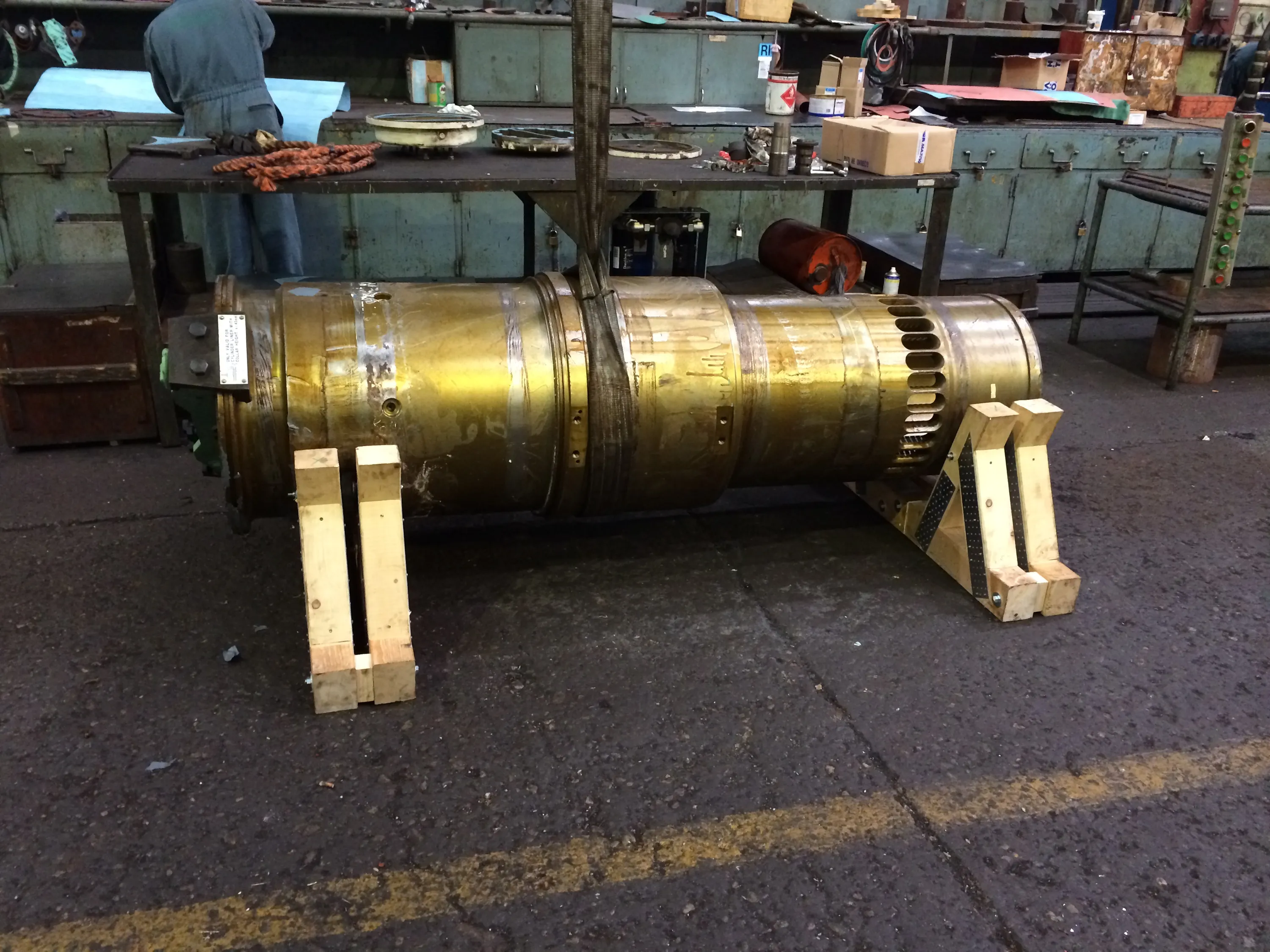 A large, golden-hued metal cylinder, possibly a machine part, rests on two light wooden supports on a dark, textured floor with a yellow line. In the background, a person in blue overalls works at a cluttered workbench, backed by teal metal cabinets. The industrial space is lit by overhead artificial light, reflecting off the cylinder's surface.