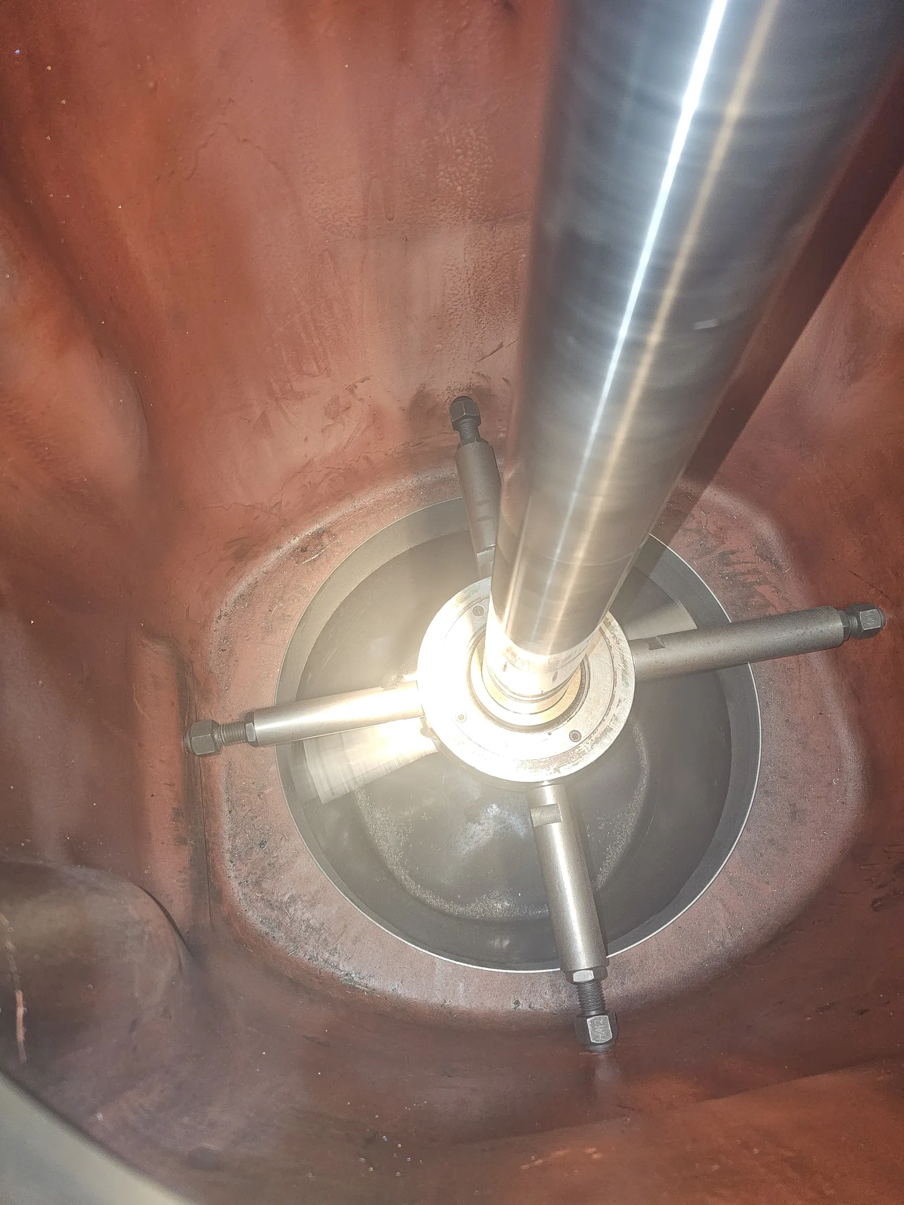 A top-down view inside a large, reddish-brown industrial tank or vessel. A prominent, reflective metal shaft runs vertically down the center, secured near the bottom by a circular metal flange with four radial arms bolted to the tank's inner wall. The inner surface of the tank is a textured, reddish-brown, showing signs of wear and some grime. The bottom of the tank appears dark and circular, with scattered light reflections or debris. Bright light from above illuminates the scene, creating strong reflections on the shaft and a bright glow around the central support structure.