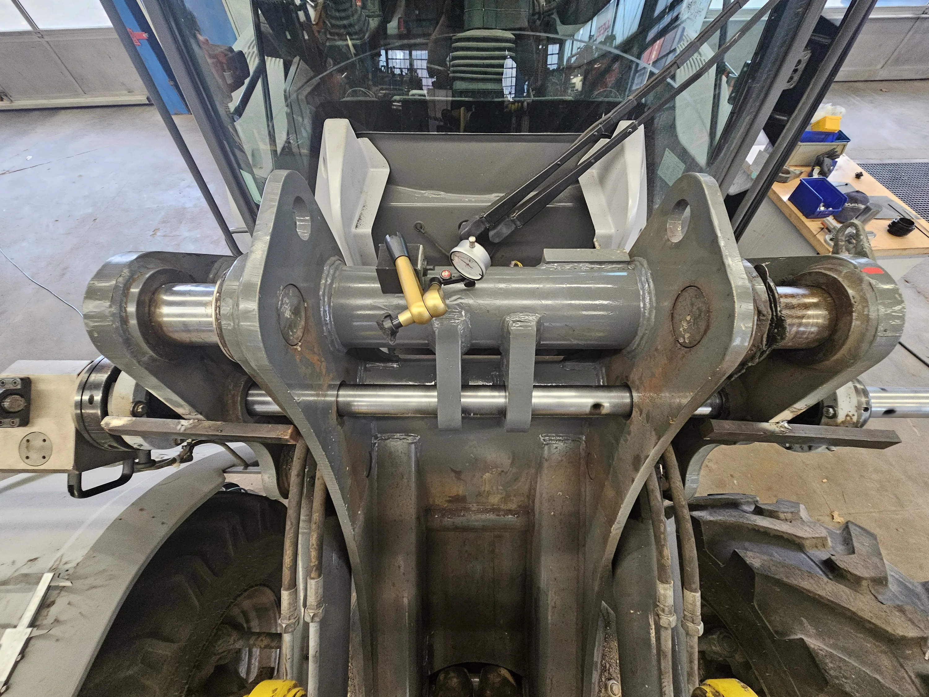 A high-angle view captures the front attachment mechanism of heavy machinery, likely a tractor, within a workshop. The robust grey metal structure features large pins and a central hydraulic cylinder, to which a dial indicator gauge and a brass-colored fitting are attached. Reflections in the machinery's windshield show the workshop interior with windows. To the right, a wooden workbench holds blue and yellow bins, tools, and a black cap. On the left, a large, dark-treaded tire and a white fender are visible against a light grey concrete floor. The scene is brightly and uniformly lit.