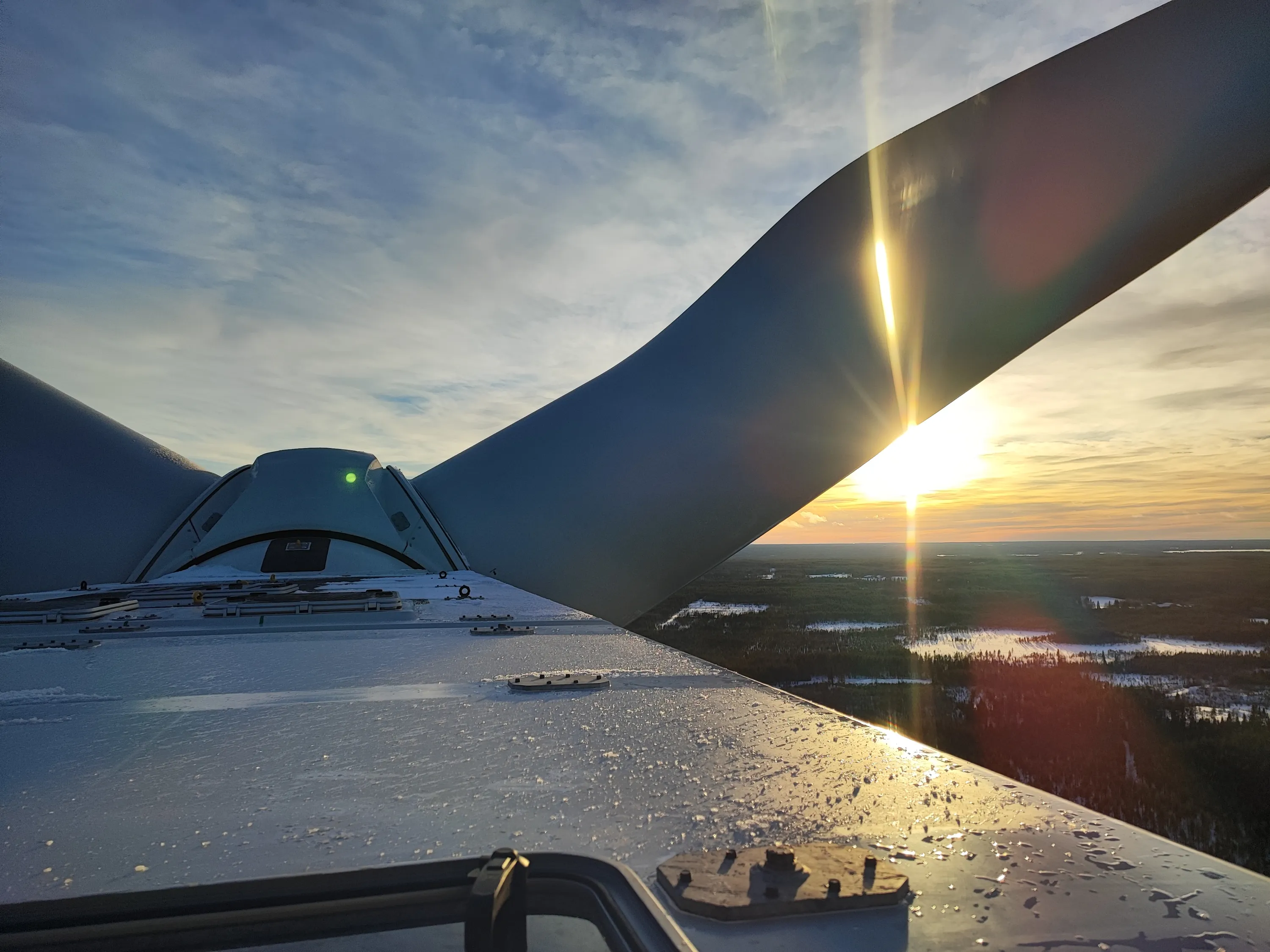 An aerial perspective from a wind turbine reveals its frosted top surface and two massive blades. The setting sun, positioned behind the right blade, emits a brilliant golden light and lens flare, illuminating the icy foreground and parts of the turbine. A distinct green light glows from the turbine's central hub. Below, a sprawling winter landscape of snow-covered forests and dark lakes extends to the distant horizon, beneath a sky painted with warm orange, yellow, and blue hues and wispy clouds.