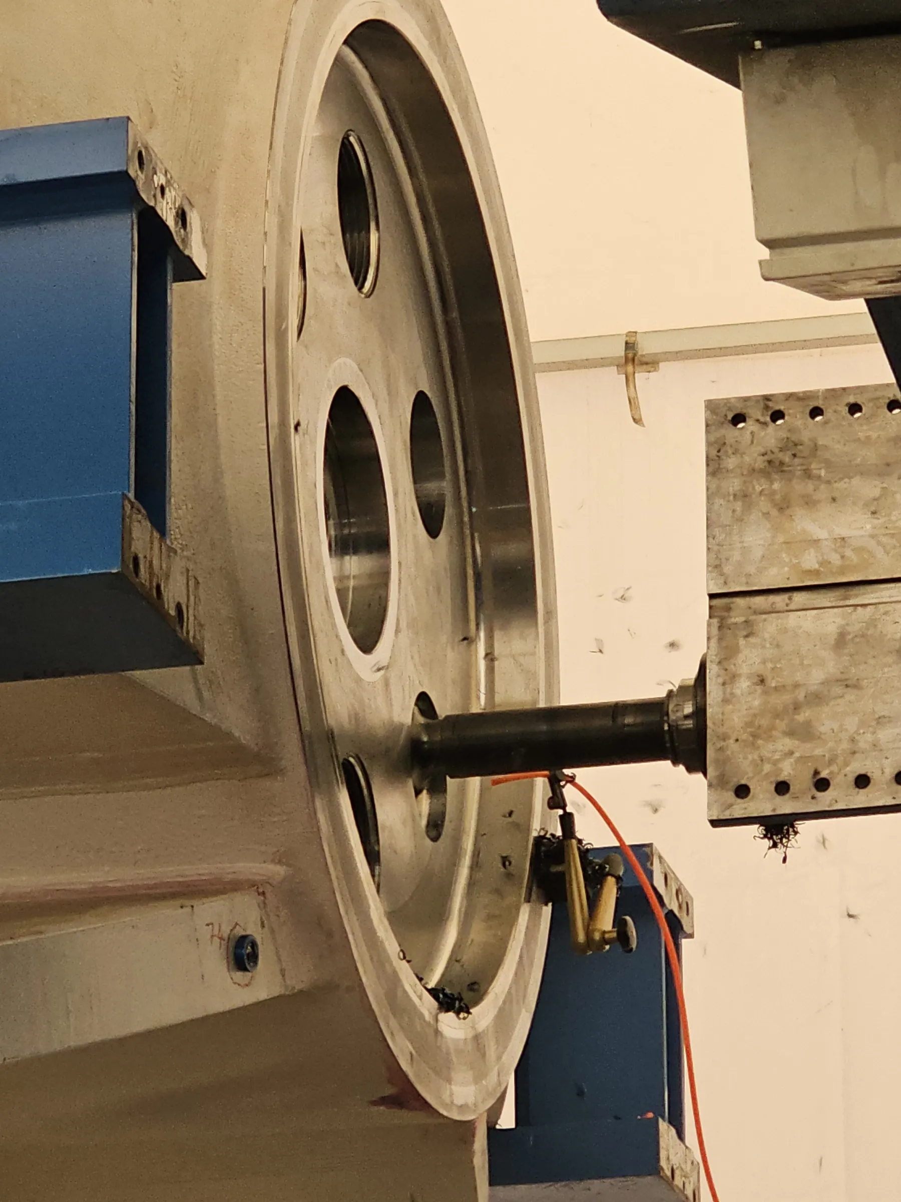 A large, silver-toned metal wheel with multiple circular openings is being machined by a black cylindrical tool. Metal shavings accumulate near the tool and in the wheel's lower groove. An orange cable connects to a small brass-colored component near the work area. A blue metal structure supports the wheel on the left, with a light beige wall in the background.