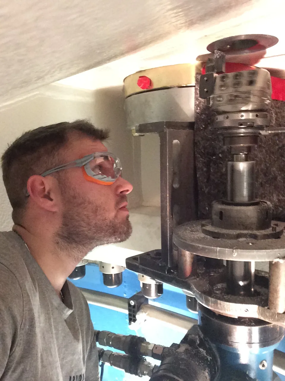 A man with a beard and a grey t-shirt wears clear safety glasses with grey and orange accents, looking up intently at complex industrial machinery. The machine features various metal components, including a rotating head and shafts, covered in fine debris. A light-colored ceiling is visible above, and a blue surface with pipes and fittings is below the main machinery. The scene is brightly lit.